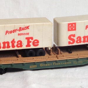 以下が含まれることがあります： 赤い文字で「Piggy-Back Service Santa Fe」と書かれた2つのクリーム色のコンテナを搭載した模型の貨車。貨車は茶色と緑色で、「Great Northern」の文字が側面に印刷されています。