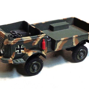 Op de afbeelding: Een speelgoed militaire vrachtwagen met een camouflagekleurschema. De vrachtwagen heeft een laadbak en een rode brandblusser aan de zijkant. De vrachtwagen heeft een zwart hakenkruissymbool aan de zijkant.