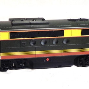 May include: A model train locomotive in dark green, yellow, and red. The train has black wheels and a detailed design with rectangular windows and circular vents. The top of the train has a series of small, rectangular features.