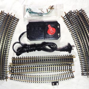 May include: A model railway set featuring curved and straight tracks, a black power supply with a red control knob, and small connectors. The tracks are black with gold-coloured rails.
