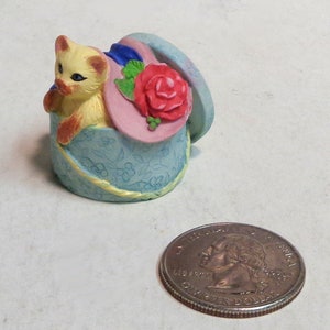 May include: A small ceramic figurine of a yellow cat wearing a pink hat with a red rose and green leaves. The cat is sitting inside a blue and white floral box with a yellow ribbon.