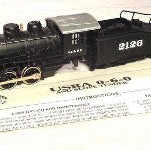 May include: A black HO scale model steam locomotive with a silver roof and the number 2126 on the tender. The train is positioned on an instruction sheet labeled USRA 0-6-0 AND SLOPE TENDER, featuring Bachmann branding and troubleshooting tips.