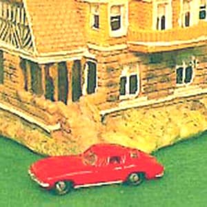 May include: A red toy car parked in front of a brown and white toy house. The house has a white balcony and multiple windows.