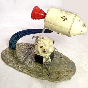 May include: A vintage model of a space shuttle and lunar lander. The shuttle is off-white with black accents and a red engine. The lander is cream-coloured with black details, set on a textured, grey base. A blue arc connects the two.