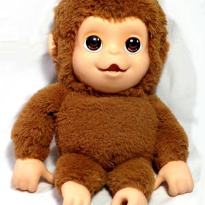 May include: A plush monkey doll with brown synthetic fur covering its head, torso, and limbs. The doll features a smooth, light-colored vinyl face with large, dark brown eyes, a small nose, and an open mouth. The hands and feet are also vinyl.