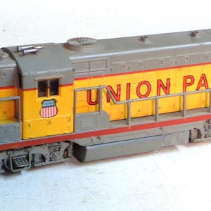 Może przedstawiać: Szary i żółty model lokomotywy kolejowej z napisem "UNION PACIFIC" w kolorze czerwonym na boku. Lokomotywa ma czerwony pasek wzdłuż dolnej części i małą amerykańską flagę z boku.