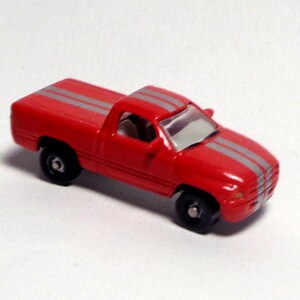 May include: A small, red toy pickup truck with two gray stripes running down the hood and roof. The truck has black wheels and a gray windshield. The toy is a miniature replica of a full-size pickup truck.
