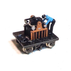 May include: A small, black and brown toy train engine with a blue and black toy figure of a person working on it. The engine is on a black platform with two wheels on each side.
