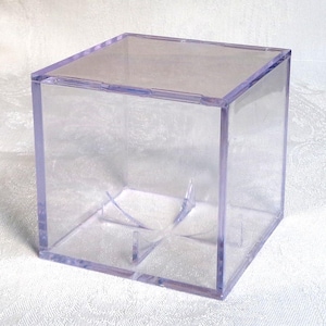 May include: A clear, square display box with a purple-tinted frame. The box has a transparent design, allowing full visibility of the contents. Interior supports are visible at the base, providing stability. The box is designed to showcase items.