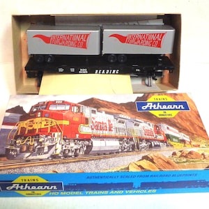Puede incluir: Una caja para un set de tren modelo de Athearn. La caja muestra una locomotora Santa Fe roja, blanca y gris tirando de un tren. La caja también muestra dos vagones de plataforma con remolques de International Forwarding Co. La caja es azul y blanca con el texto "TRAINS Athearn IN MINIATURE" y "AUTHENTICALLY SCALED FROM RAILROAD BLUEPRINTS HO MODEL TRAINS AND VEHICLES".