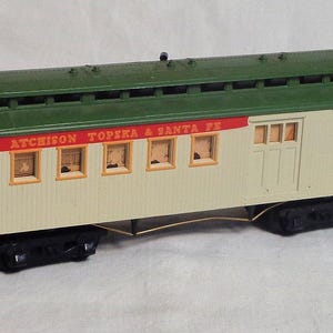 May include: A model train passenger car with a cream-colored body, green roof, and black undercarriage. The car has windows and the words "Atchison Topeka & Santa Fe" in red lettering. The model train car is a vintage collectible.