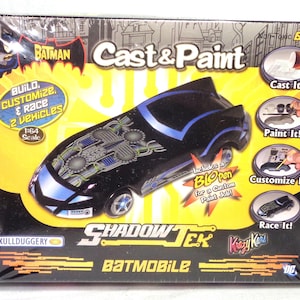 May include: A Batman Cast & Paint ShadowTek Batmobile 1:64 scale model kit box. The packaging features a black and blue toy car, yellow background, and text including Build, Customize, & Race 2 Vehicles, Includes a BLOpen for a Custom Paint Job!
