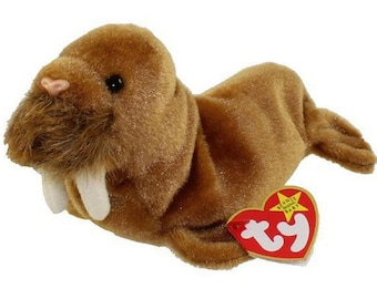 May include: A brown plush walrus toy with large white tusks and a pink nose. The stuffed animal has a soft, textured appearance and a red heart-shaped tag with the words "Beanie Baby" on it.
