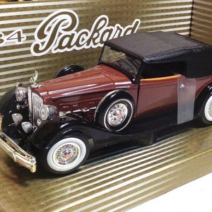 May include: A 1934 Packard model car in brown and black with a black convertible top. The car is displayed in a box with a gold and white striped background.