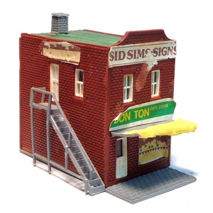 May include: A red brick building with a green roof and a yellow awning. The building has a sign that says "Sid Sims Signs" and another sign that says "Don Ton Dept. Store". There is a fire escape on the side of the building.