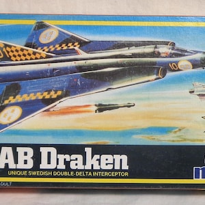 Puede incluir: Una maqueta de un avión de combate Saab Draken azul y amarillo. El arte de la caja muestra el avión en vuelo con un patrón de cuadros amarillo y negro. La caja está etiquetada como "SAAB Draken" y "Unique Swedish Double-Delta Interceptor". La caja también está etiquetada como "1/72 Model Kit" y "Modele Reduit" y tiene el logotipo de MPC.