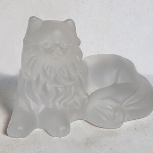 Puede incluir: Una figura de gato de vidrio esmerilado en posición reclinada. El gato tiene detalles de pelaje y está acostado con las patas delanteras extendidas. La figura es de color claro y translúcido.