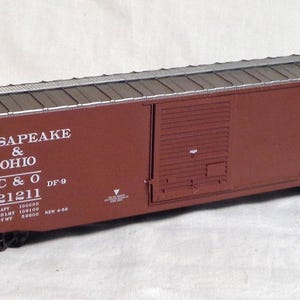 May include: A model of a Chesapeake & Ohio C&O 21211 freight car. The car is painted maroon with white lettering and a silver roof. The text "CHESAPEAKE & OHIO" and "C & O" are visible. The car has black wheels and is on a white surface.