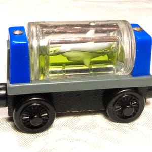 May include: A toy train car with a clear, cylindrical container holding a white shark figurine in green liquid. The train car has blue and gray accents, black wheels, and is made of wood.