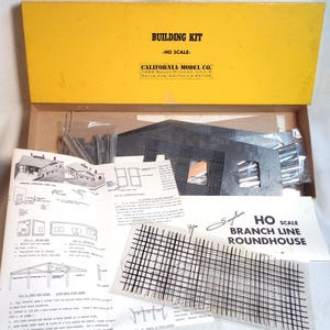 Op de afbeelding: Een open doos met een HO-schaal modelbouwkit. Het gele deksel leest "BUILDING KIT - HO SCALE" en "CALIFORNIA MODEL CO." Binnenin zitten bouwcomponenten, instructies en een doorzichtige plastic plaat met een rasterpatroon.