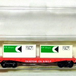 Könnte beinhalten: Ein Modell-Eisenbahnwagen in einer transparenten Kunststoffbox. Der Wagen ist rot mit schwarzen Rädern und trägt zwei weiße Container mit grünen "CP Ships"-Logos und schwarzen Pfeildesigns. Die Wagennummer ist CN 657036.