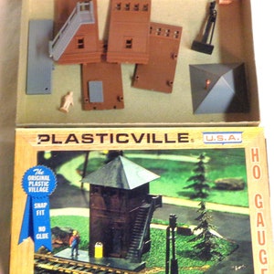 May include: A plastic model kit of an HO gauge signal box. The kit includes a brown tower with a grey roof, a ladder, and a signal light. The box also includes the text "PLASTICVILLE. U.S.A. HO GAUGE" and "The Original Plastic Village Snap Fit No Glue Switch Tower 2619-100".