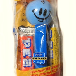 May include: A blue and white PEZ dispenser with a smiling face. The dispenser is packaged in a red, yellow, and green plastic bag. The bag has the text "Candy & Dispenser" and "Visit our website www.pez.com". The bag also has the text "Candy Net Wt: .58 oz. (16.4g)".