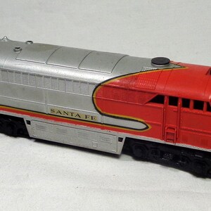May include: A model diesel locomotive train engine featuring a silver body with a bright red nose and cab section. The side displays the text SANTA FE in black lettering, accented by thin yellow and red horizontal stripes along the lower midsection.