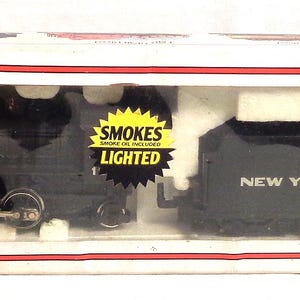 May include: A black model train set in a white box with a red border. The train is labeled "New York Central" and has a "Smokes Lighted" sticker. The box is slightly damaged.
