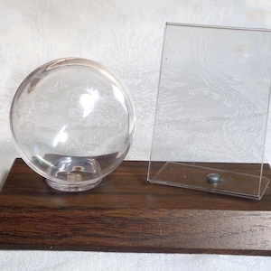 May include: A clear glass globe and a rectangular glass panel are mounted on a dark brown wooden base. The globe sits on the left, and the panel leans against the base on the right. The base has a rectangular shape.