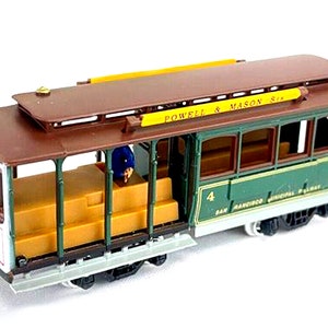 May include: A miniature cable car model with a brown roof and green sides. The model has the words "POWELL & MASON Sta." and "SAN FRANCISCO MUNICIPAL RAILWAY" printed on it. The cable car has a driver and yellow seating.