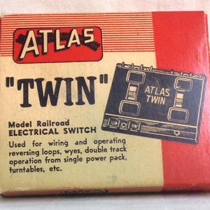 Może przedstawiać: Czerwono-białe pudełko z nadrukowanymi słowami "ATLAS" i "TWIN". Pudełko zawiera model kolejowy przełącznik elektryczny. Tekst na pudełku brzmi: "Służy do okablowania i obsługi pętli odwracających, rozjazdów, podwójnego toru, obsługi z jednego zasilacza, stołów obrotowych itp."