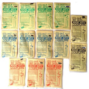 May include: A collection of vintage HO scale Magne-Matic Couplers in various packages. The packages are primarily cream-colored with green, red, and blue text. Each package displays the coupler number and includes the text "Two Pair with draft gear".