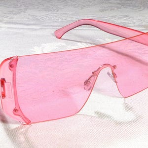 May include: Pink sunglasses with a shield-style design. The frames and lenses are a solid, translucent pink. The sunglasses have a curved brow bar and straight temples. The lenses are a large, rectangular shape.