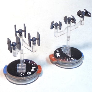 May include: Two Star Wars X-Wing TIE Fighter miniatures on clear plastic stands. The miniatures are painted black and gray. The stands are attached to circular bases with numbers 1, 2, and 3 printed on them.