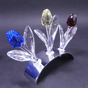 Puede incluir: Tres tulipanes de cristal en azul, amarillo y rojo sobre un soporte plateado. Los tulipanes están hechos de cristal transparente con acentos de color.