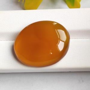 Op de afbeelding: Een gepolijste, ovaalvormige edelsteen in een warme amberkleur. De steen heeft een glad, reflecterend oppervlak, met highlights die wijzen op een hoge mate van helderheid. De edelsteen is geplaatst tegen een witte achtergrond, met een vleugje gele bloemelementen.