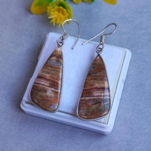 Könnte beinhalten: Ein Paar Ohrringe mit silberfarbenem Metallrahmen. Die Ohrringe haben eine Tropfenform mit einem Naturstein-Design in Braun-, Rot- und Cremetönen. Die Ohrringe werden auf einer weißen Oberfläche präsentiert.