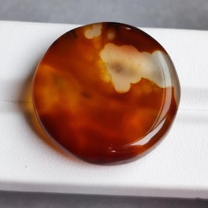 Könnte beinhalten: Ein polierter, runder Edelstein mit einer warmen, erdigen Farbpalette. Der Stein weist eine Mischung aus tiefem Rotbraun und Bernsteintönen auf, mit einem durchscheinenden, wolkenartigen Muster in Creme. Die Oberfläche ist glatt und reflektierend, was auf eine hochwertige Verarbeitung hindeutet.