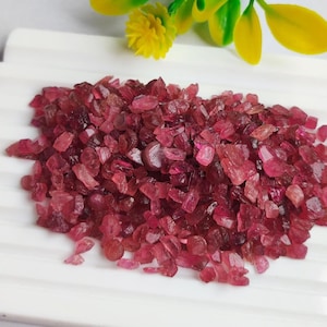 Könnte beinhalten: Ein Haufen kleiner, grob geschnittener, rubinroter Edelsteine. Die Edelsteine ​​liegen auf einer weißen, geriffelten Oberfläche, mit gelben und grünen floralen Akzenten im Hintergrund. Die Edelsteine ​​scheinen roh und unpoliert zu sein.