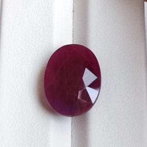 Puede incluir: Una gema de rubí facetada de forma ovalada. El rubí es de un color rojo intenso y rico con una superficie pulida. Las facetas reflejan la luz, creando un efecto brillante. La gema está sobre un fondo blanco.