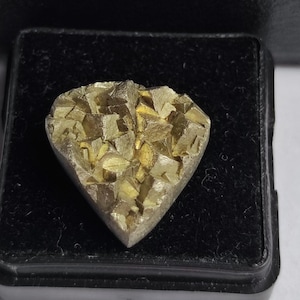 May include: A heart-shaped, metallic-looking object with a silver border and a textured, golden-yellow center. The center appears to be composed of many small, cube-like structures. The object is set against a black background.