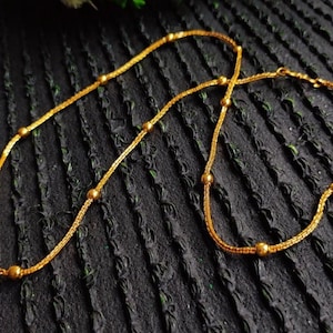 Puede incluir: Un collar de cadena en tono dorado que presenta una serie de pequeñas cuentas esféricas pulidas, espaciadas uniformemente e integradas a lo largo de la delicada cadena de eslabones texturizados, creando un patrón constante y repetitivo.