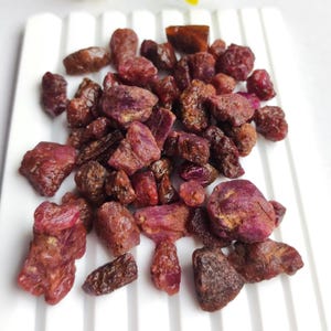 Puede incluir: Un conjunto de piedras preciosas de rubí en bruto, sin tallar, en varios tonos de rojo y púrpura. Las piedras tienen un aspecto natural y texturizado, dispuestas sobre una superficie blanca. Los rubíes son de diferentes tamaños y formas.