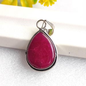 Può includere: Un ciondolo a forma di goccia con una grande pietra lucida color magenta intenso. La pietra è incastonata in una montatura in metallo color argento con un semplice anello per una catena. Il ciondolo è esposto su una superficie bianca, con fiori gialli sullo sfondo.