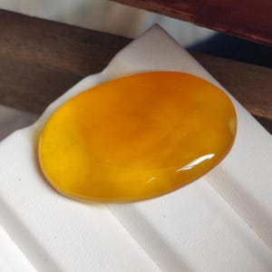 May include: A polished, oval-shaped gemstone with a vibrant, translucent orange hue. The stone is smooth and glossy, reflecting light. The gem is set against a white surface, with a wooden element in the background.