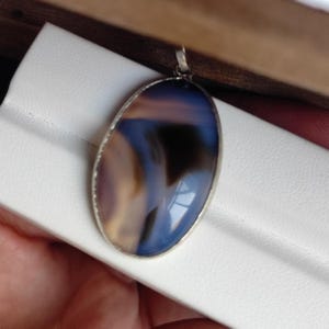 Könnte beinhalten: Ovaler Anhänger mit einem polierten, durchscheinenden Stein. Der Stein weist eine Mischung aus blauen, braunen und cremefarbenen Farben auf, die in einem silberfarbenen Metallrahmen gefasst sind. Der Anhänger hat eine kleine Schlaufe für eine Kette.
