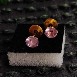 May include: A pair of stud earrings featuring round, faceted light pink stones set in gold-toned metal. Each earring has a circular gold-toned backing and is displayed on a small, rectangular white foam block with a black top surface.