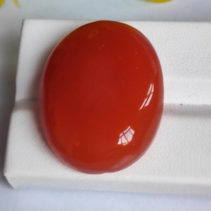 May include: A polished, oval-shaped gemstone in a vibrant shade of red. The gem has a smooth, glossy surface, reflecting light. The stone is set against a white background, highlighting its color and shape. This gemstone could be used in jewelry.