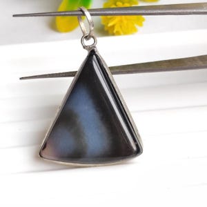 Puede incluir: Un colgante triangular con un marco y una fianza de color plateado. La piedra del colgante tiene una superficie oscura y brillante con un patrón arremolinado de azul y gris. El colgante está sujeto por unas pinzas.